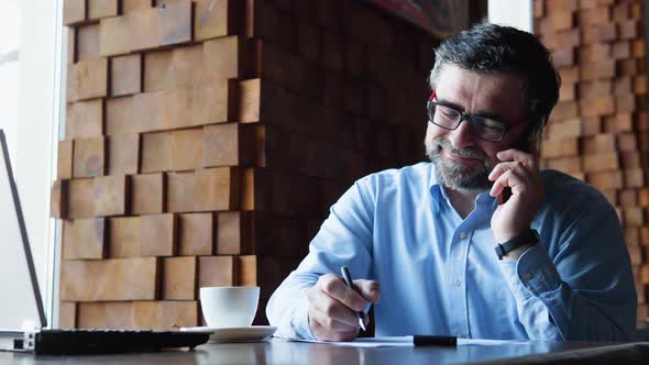 Close Up of Senior Businessman Talking on the Mobile Phone Looking at Laptop in Cafe