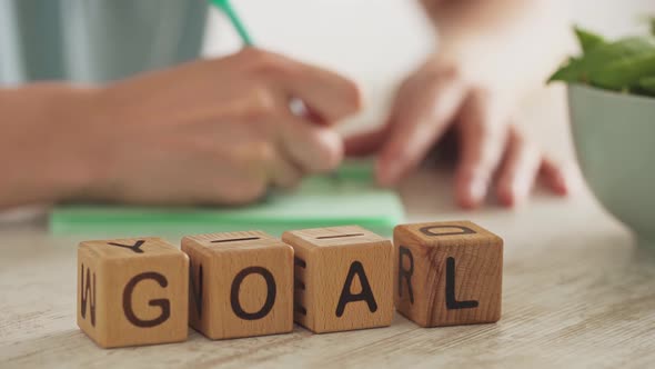 The Word Goal Is Assembled From Wooden Cubes. In The Background, A Female Hand Writes Down The Goal