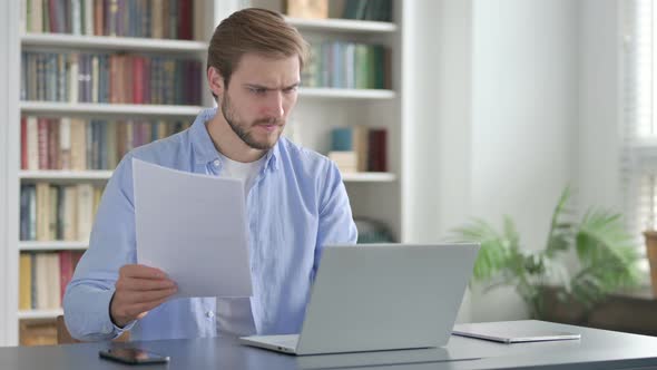 Upset Man Reading Documents While Using Laptop