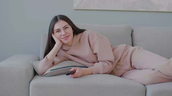Middle frame of young woman with glasses lying on sofa in good mood and changing TV channels
