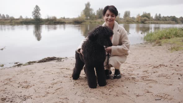 A Smiling Young Woman in a Stylish White Coat is Squatting on the River Bank and Hugging a Large