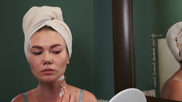 Young Woman Using Jade Facial Roller for Neck Massage Sitting By the Bathroom Mirror Slow Motion