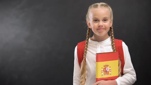 Smiling Schoolgirl Holding Spanish Language Book Against Blackboard Background