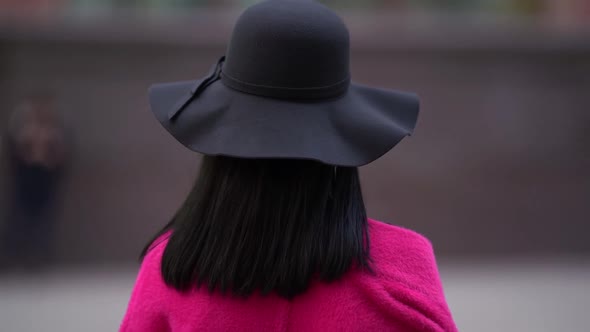 Close-up Portrait of a Stylish Brunette Woman in a Pink Coat and Black Hat Walking on a Blurry