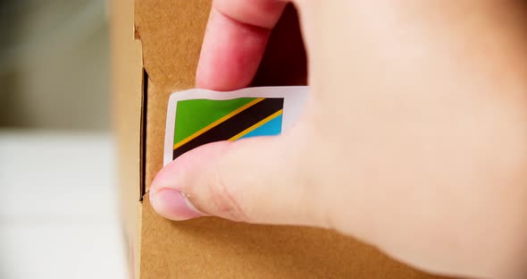 Hands applying MADE IN TANZANIA flag label on a shipping cardboard box with products. Close up shot