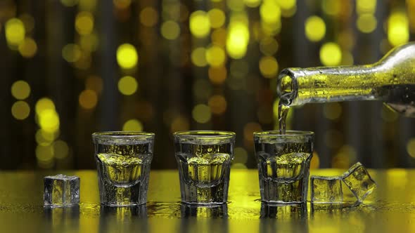 Barman Pour Frozen Vodka From Bottle Into Shot Glass. Ice Cubes Against Shiny Gold Party Background