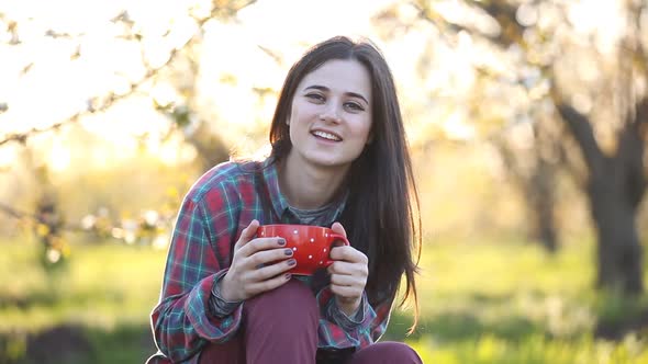 Brunet girl in shirt with a cup next to an apple tree garden.