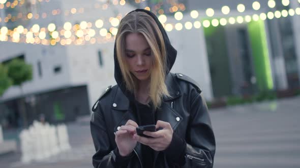 Young Girl Uses a Smartphone Near a Modern Residential Area Evening in the City Blurry Lights on