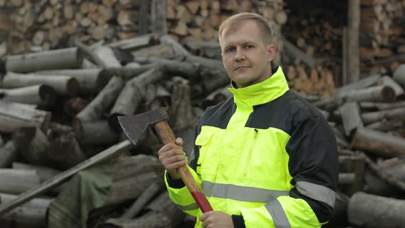 Man Woodcutter Lumberjack with Small Axe. Sawn Logs, Firewood Background. Asks To Follow Him