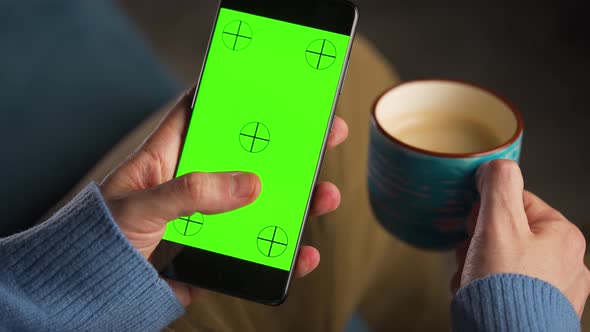 Man Using Smartphone with Green Mockup Screen in Vertical Mode and Drinking Coffee