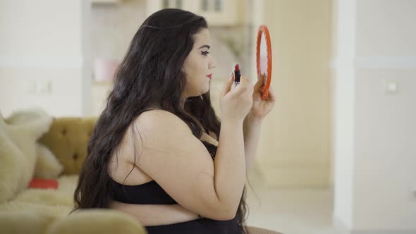 Portrait of Attractive Young Plus Size Girl Doing Make-up Looking in the Mirror. Side View of Plump