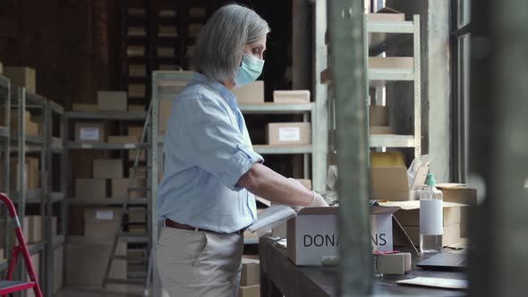 Mature Female Warehouse Worker Wearing Mask Packing Donations Box