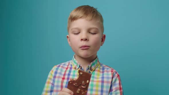 Happy Boy Eating Chocolate Bar Holding in Hands Smiling at Camera Standing on Blue Background