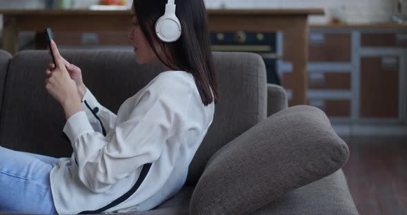 Happy Asian Woman Listening Songs Using Headphones Holding Smartphone Touching Screen Lying on Cozy