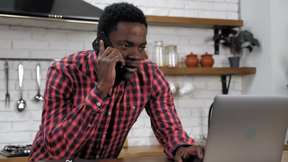 African American Man Uses Laptop Talking on Mobile Phone Standing in Kitchen