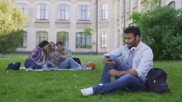 Young Hispanic Male Student Sitting on Grass and Scrolling on Cellphone, Relax