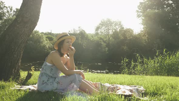 Lady eating apple sitting on meadow in park by the river and enjoying warm summer holidays