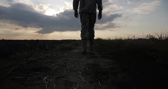 A Soldier in Military Uniform Walks Across a Black Burnt Field at Sunset