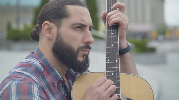 Side View of Concentrated Man Playing Guitar Outdoors. Portrait of Bearded Young Caucasian Musician