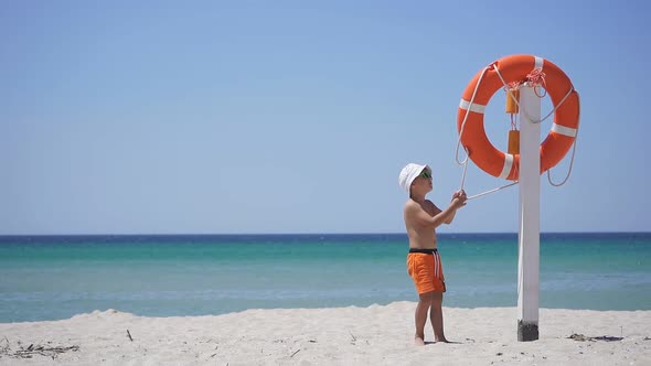 Orange Lifebuoy Hanging From a Pole on the Beach. Curious Boy on the Beach Stands Near the Lifebuoy