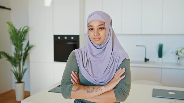 Modern Young Tattooed Arab Woman in a Lilac Hijab Stands in the Kitchen with Her Arms Folded