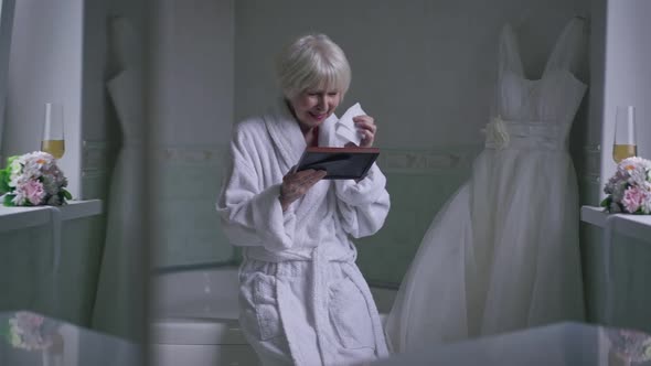 Wide Shot Crying Senior Beautiful Woman Sitting in Bathroom with Wedding Dress Looking at Photo