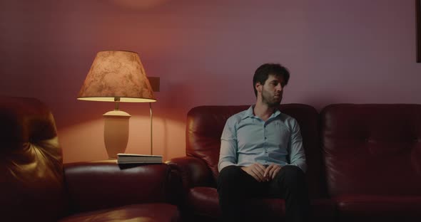 Young guy with shirt is resting sitting in the red living room of his house at night