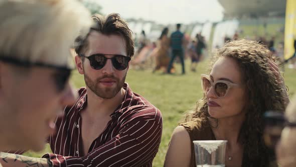 Group of friends sitting on grass together at music festival and drinking beer while chatting. Shot