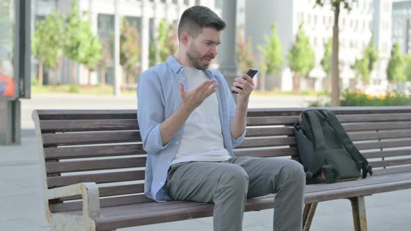 Young Man Reacting to Loss on Smartphone While Sitting on Bench