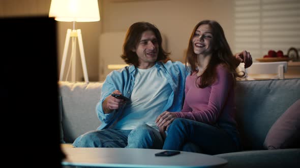 Happy Young Couple Sitting Down on Couch at Home Embracing and Choosing Program on Tv Slow Motion