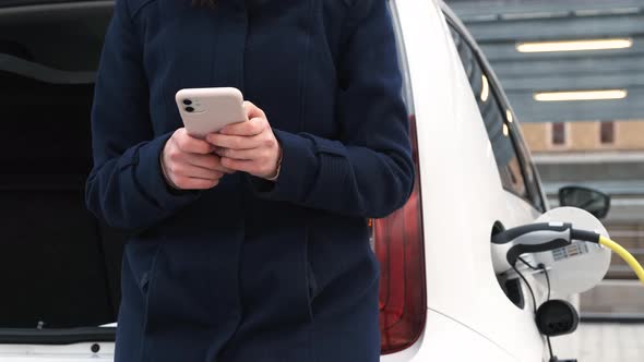 Woman Standing Near Trunk Using Her Mobile Phone and Waiting for Charging of Electric Car