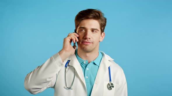 Young Doctor Man in White Medical Coat Talking on Mobile Phone on Blue Background. Doc Have