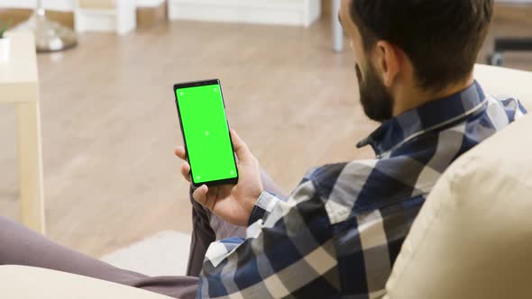 Young Man Siting on the Couch Looking at Smartphone with Green Screen on It