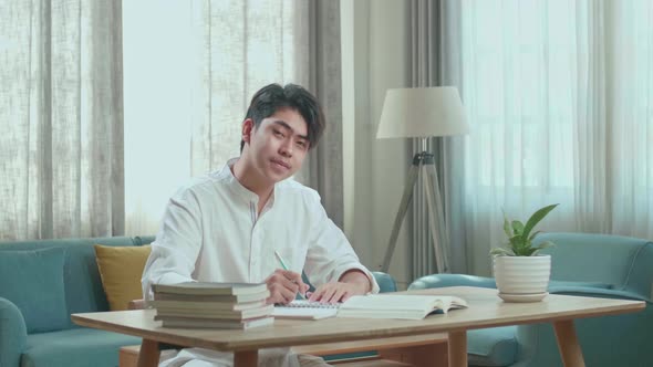 Asian Man Student Look At Camera And Smile While Writing In The Notebook On The Table At Home