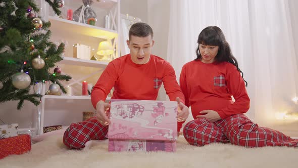 Wide Shot Portrait of Excited Caucasian Man and Pregnant Woman Opening Christmas Present and Kissing