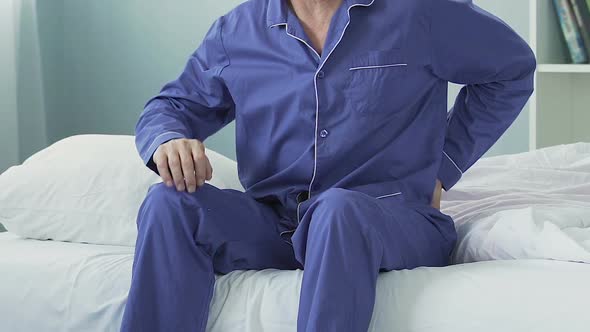 Elderly man sitting on bed edge, stretching and having sudden lower back pain