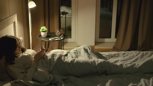 Girl in Glasses Lies in Bed at Night Covered with a Blanket Uses a Mobile Application