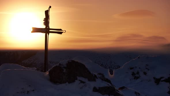 Christian cross on top of a mountain. Clouds spill over the snowy hills in the glow of the sunset