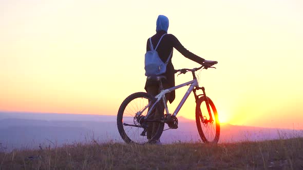 Muslim Woman in a Hijab with a Backpack with a Bicycle From the Mountain Looks at a Sunsetslow Mo