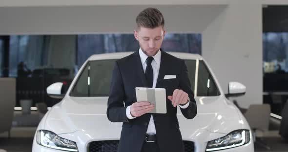 Succesful Young Car Dealer Standing in Front of White Automobile with Tablet, Looking at Camera and