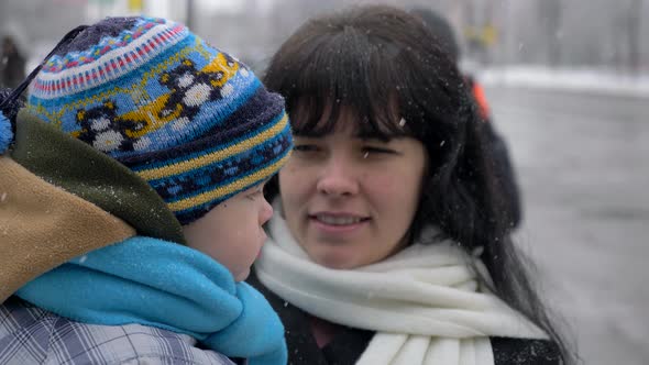 Beautiful Mother Hold Her Child On Hands. Snowflakes Falling. People With Children On Street