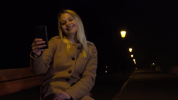 A Young Beautiful Woman Sits on a Bench and Takes Selfies with Smartphone in an Urban Area at Night