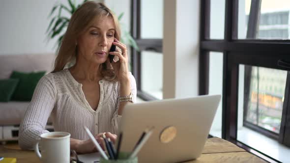 Woman Talking on Phone and Using Laptop at Table in Apartment Room