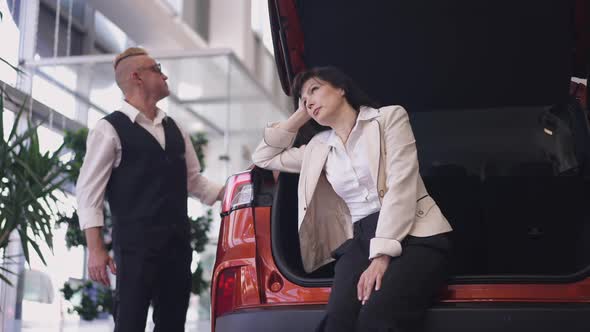 Portrait of Bored Woman Sitting in Car Trunk Sighing As Man Choosing New Vehicle in Dealership in