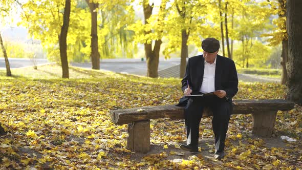 Elegant Senior Man Sitting on Bench in Park Thinking When Writing in Notebook