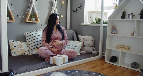 Pregnant Beautiful Woman in Pink Pajamas Sitting on the Floor Near the Christmas Tree Holding a Gift