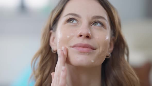 Headshot Portrait of Charming Positive Caucasian Young Slim Woman Applying Facial Moisturizer