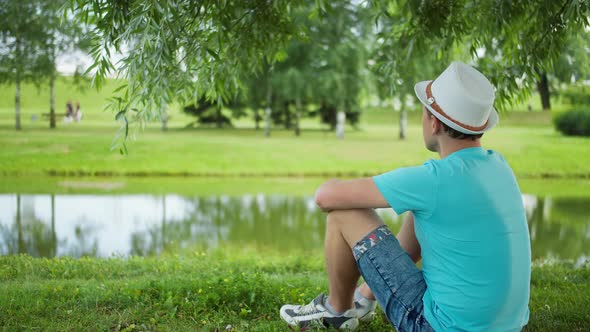 Young man in a white hat sits on the shore in a city park, back view