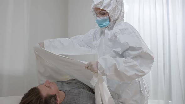 Nurse in Protective Suit Eyeglasses and Face Mask Covering Dead Man with White Sheet