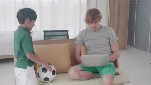 Young father working on laptop computer and son persuade play football while busy at home.